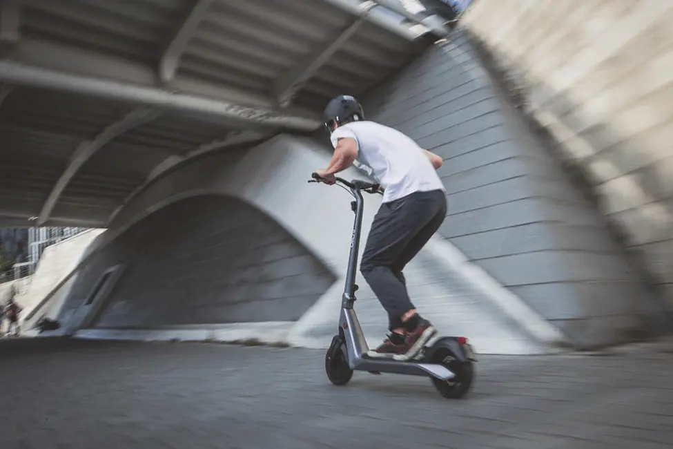 A man wearing a white shirt n black pants  and a helmet riding a scooter going under a bridge
