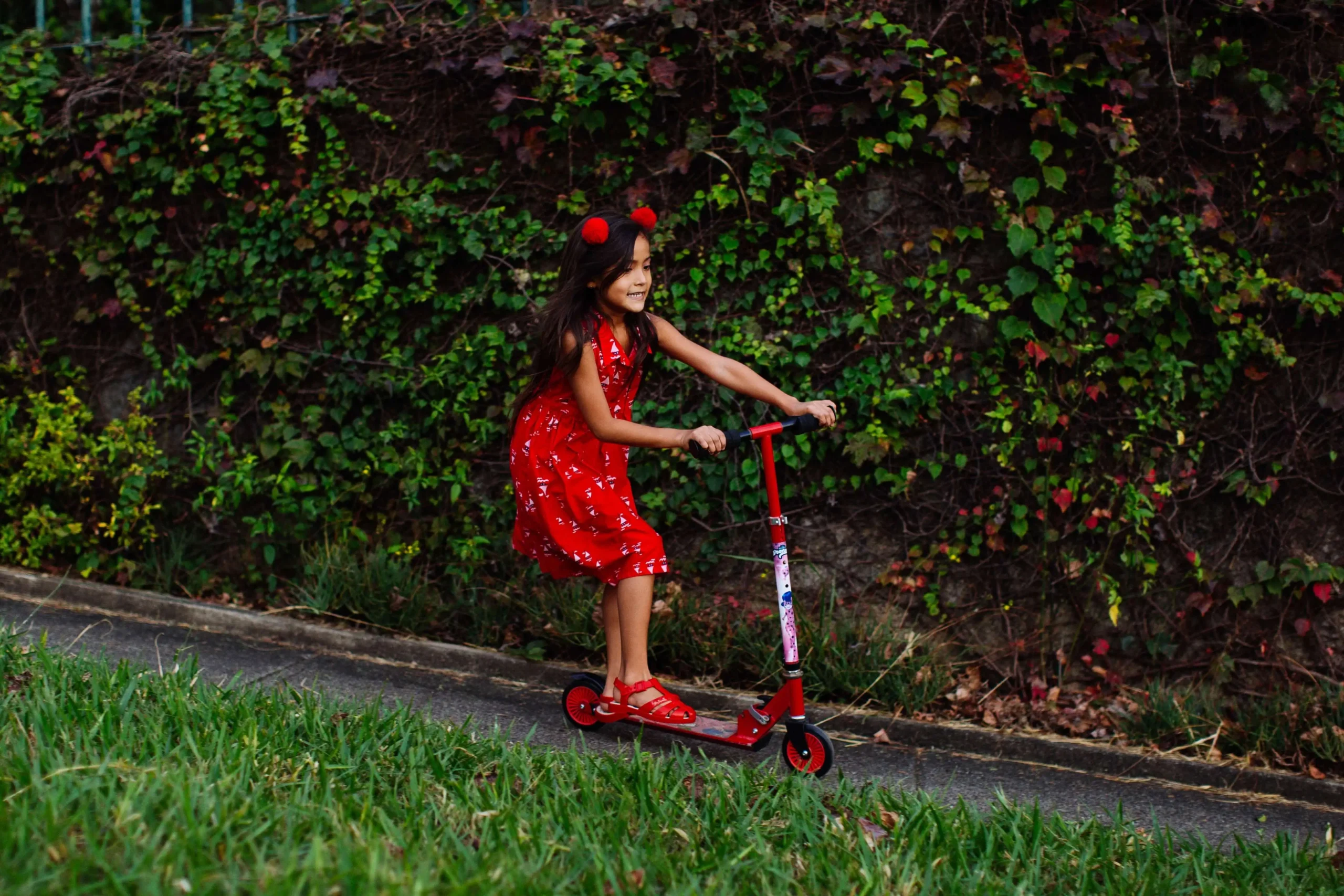 A girl in red frock riding a red scooter on the side of the grass