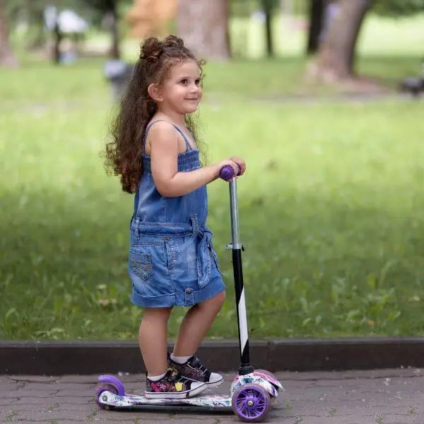 A toddler girl in blue riding a three-wheel scooter in a park