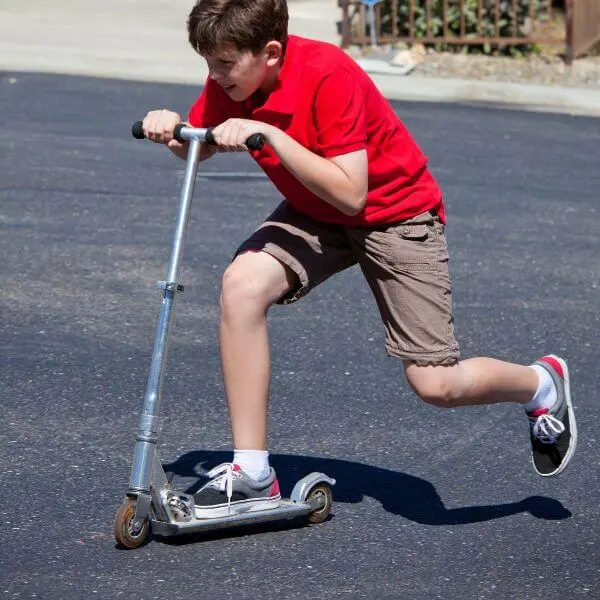 A child in a red shirt and brown shorts riding a kick scooter on the road