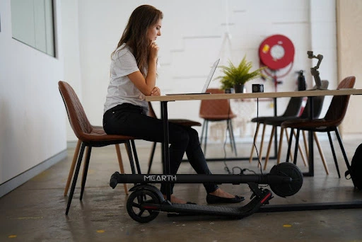 A woman looking at a laptop on a table with chairs around and a foldable scooter is rested besides her